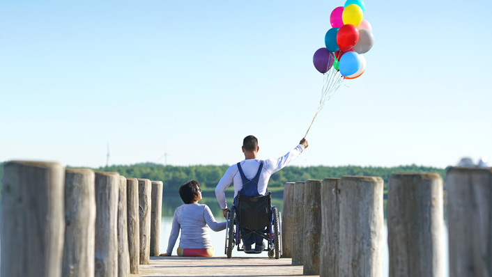 Ein Junge im Rollstuhl hält bunte Luftballons neben einem Freund auf einem Holzsteg am Wasser. - © Jenny Sturm - stock.adobe.com