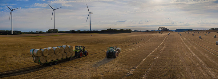 Zwei Traktoren auf einem abgeernteten Feld mit Heuballen, im Hintergrund Windräder bei teilweise bewölktem Himmel. - © Foto: snapshotfreddy - stock.adobe.com